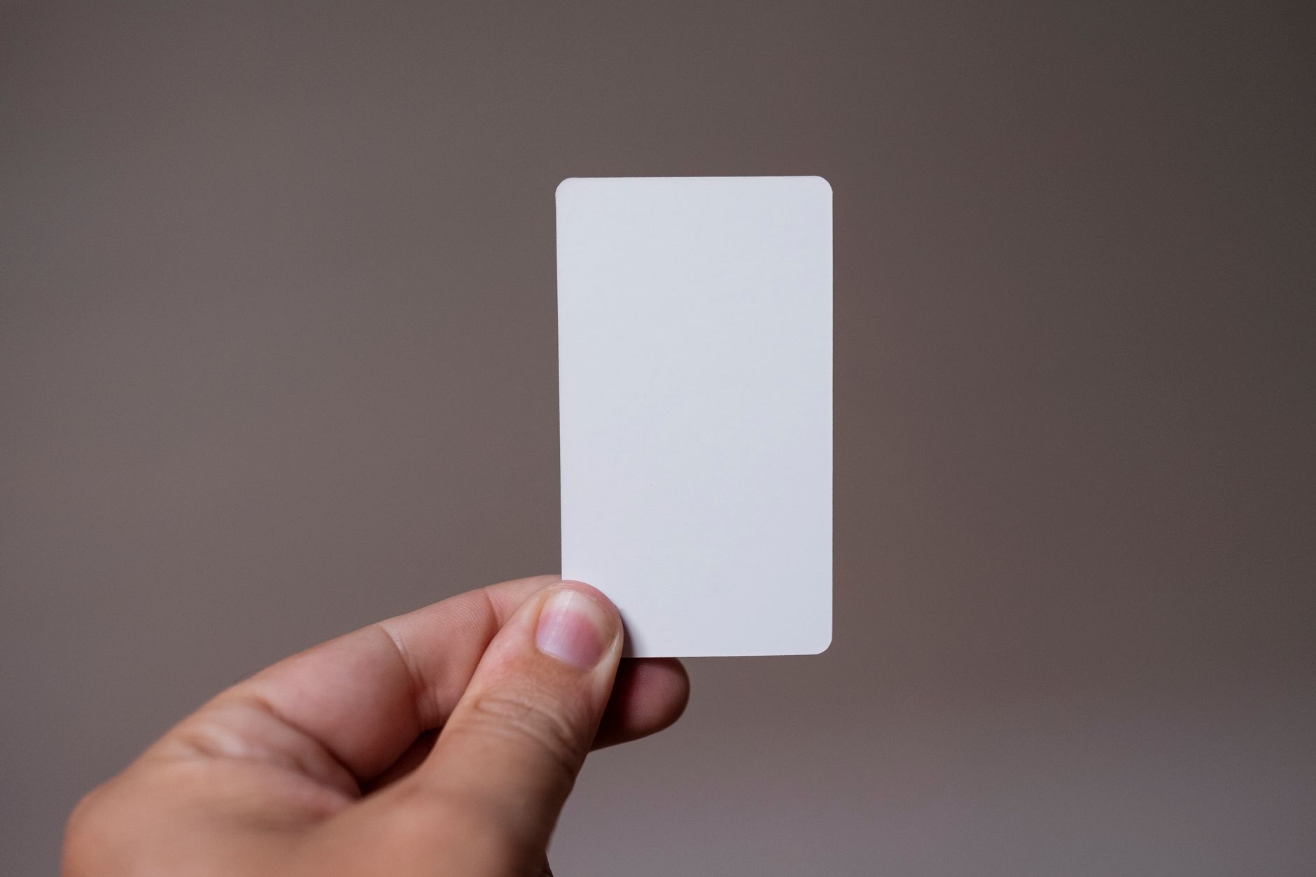 Une main tient une carte blanche vierge aux coins arrondis devant un fond gris-brun uni.