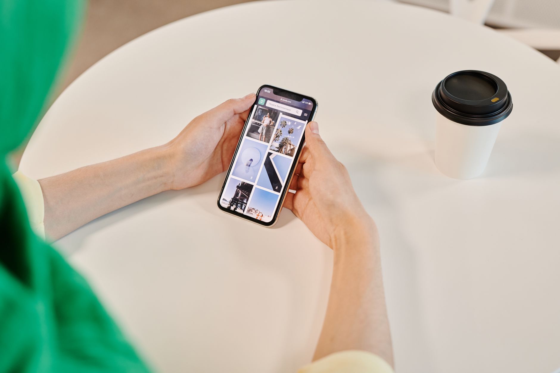 Une personne tient un smartphone affichant une grille d’images, assise à une table blanche près d’un gobelet à emporter.